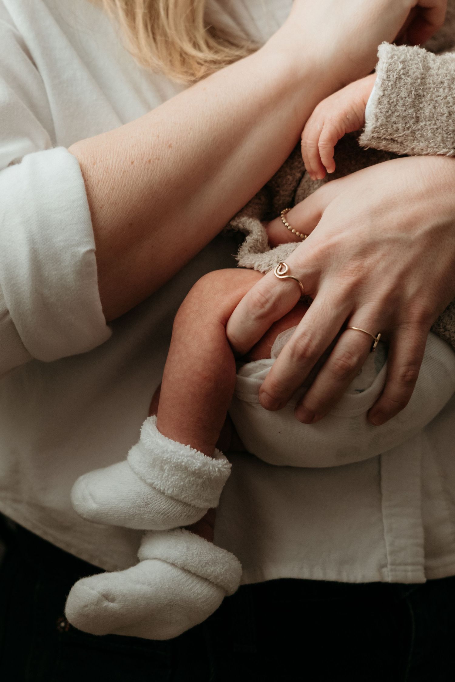 A mother cradles her newborn baby wearing white socks, her hands adorned with delicate gold and silver rings.