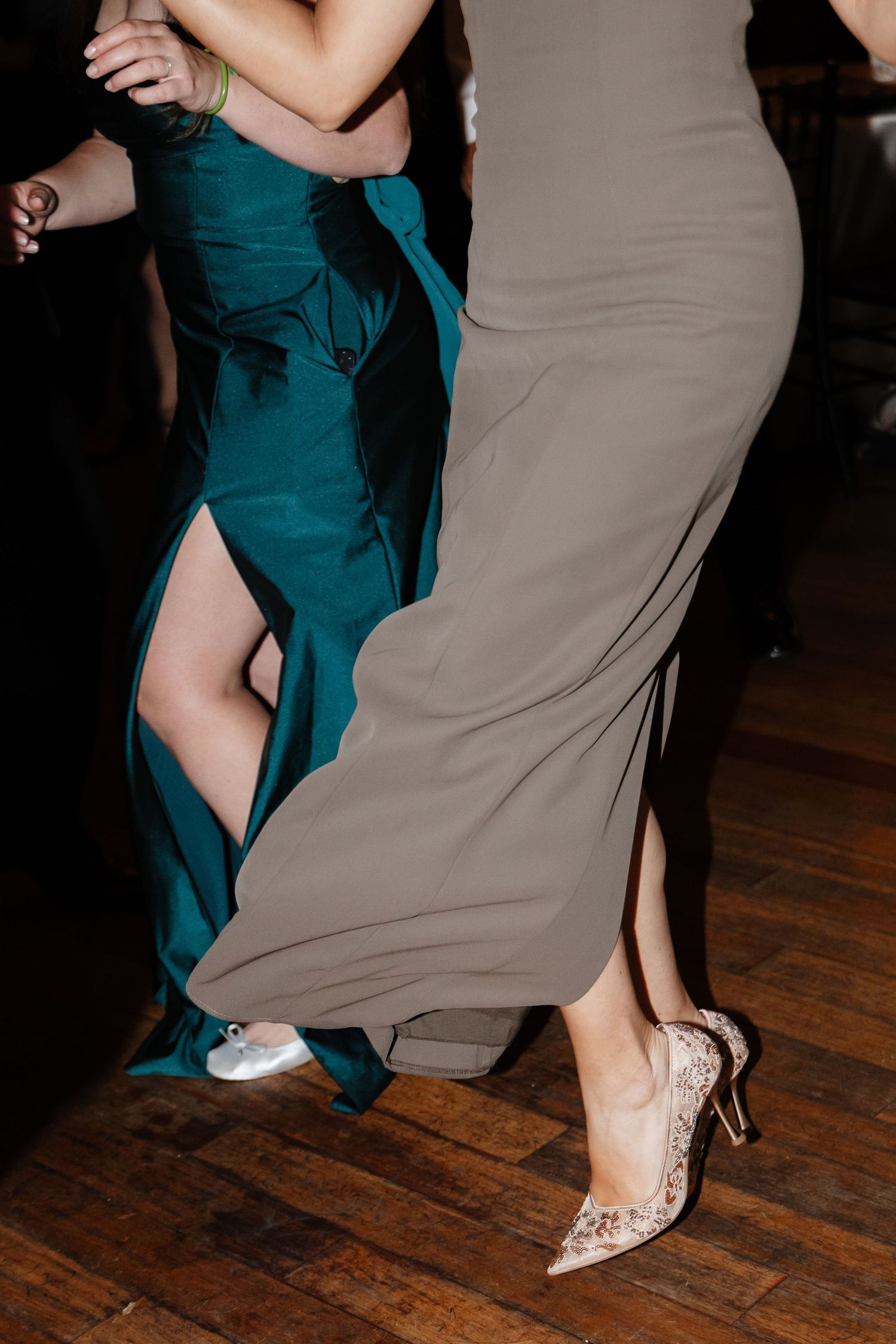 Two women dancing at a wedding reception in elegant gowns, one teal and one taupe, on a hardwood floor.