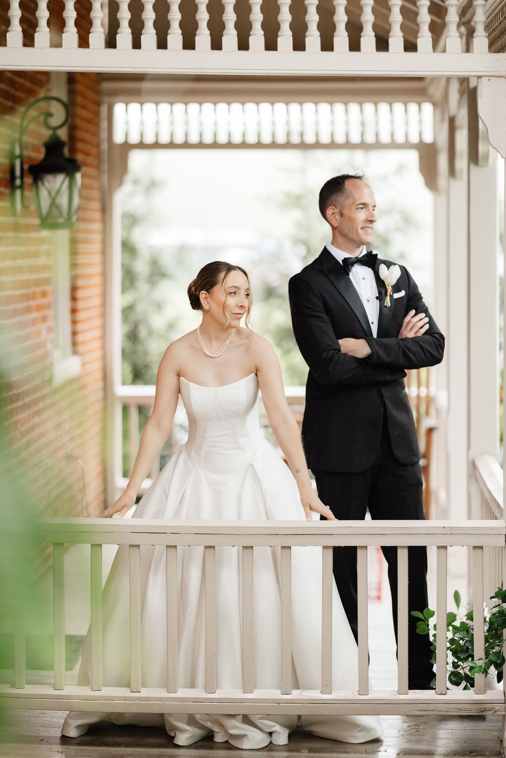 Bride and groom in formal wedding attire stand together on a porch of a brick estate venue.