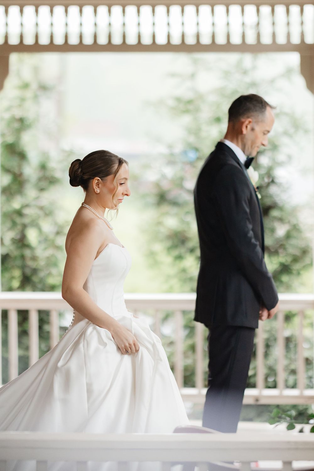 Smiling bride in strapless satin gown approaches groom in black tuxedo during outdoor first look ceremony.