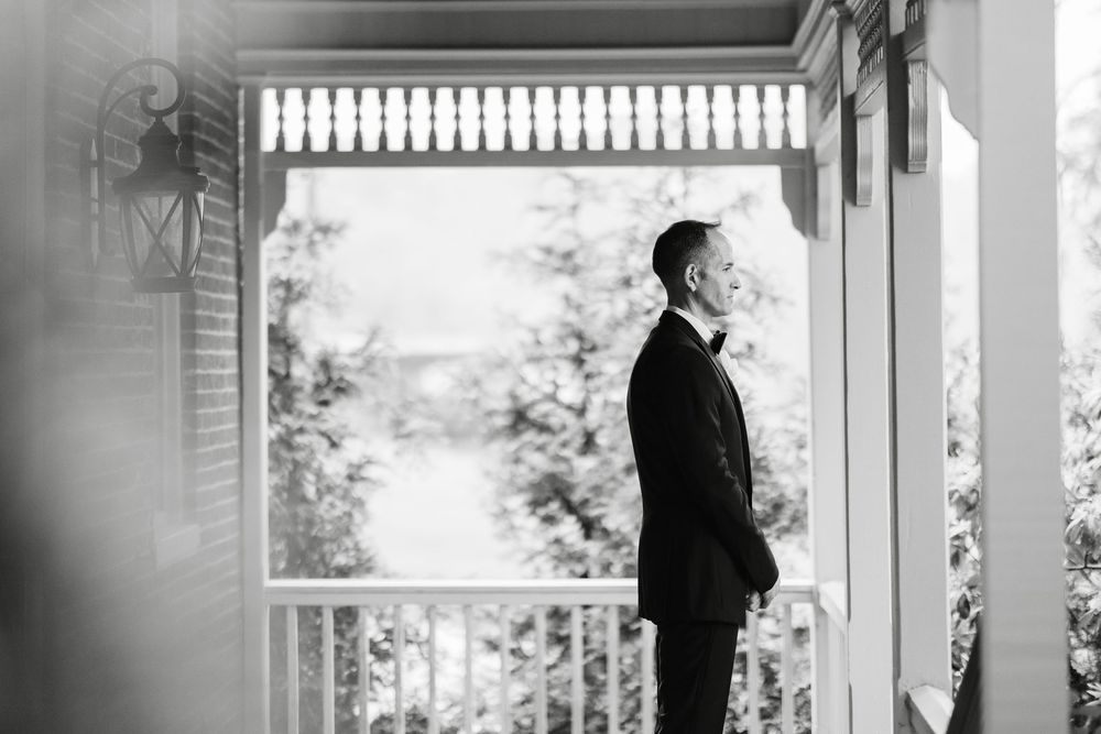 Groom in black tuxedo stands alone on white porch gazing into distance in black and white wedding photo.