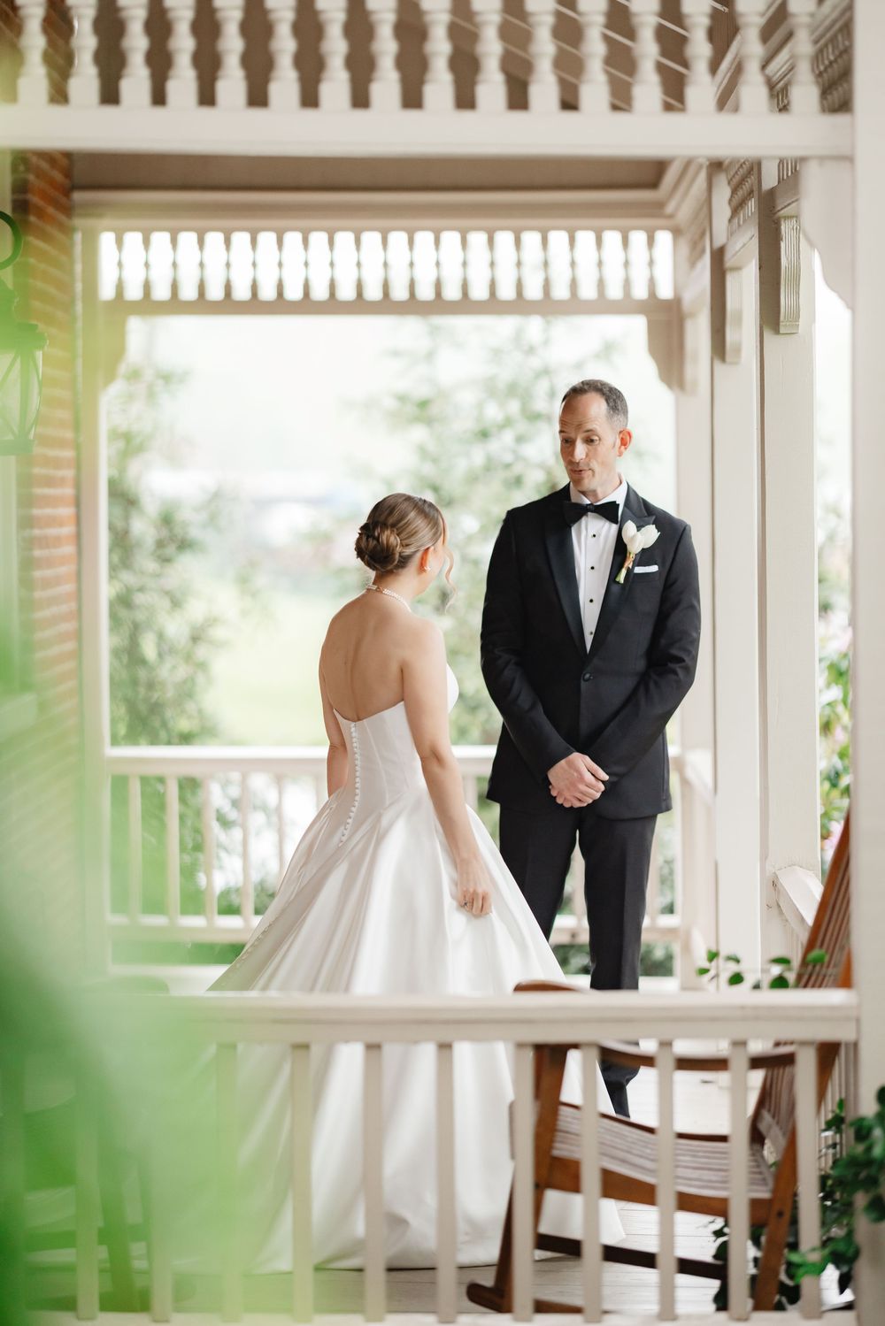 Bride in strapless white ball gown faces groom in black tuxedo during first look on elegant porch.