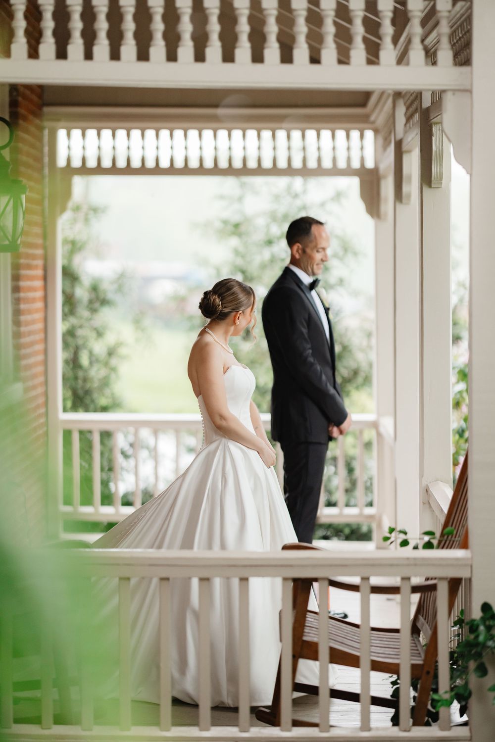 Bride and groom share a tender first look moment on a white colonial-style porch surrounded by greenery.
