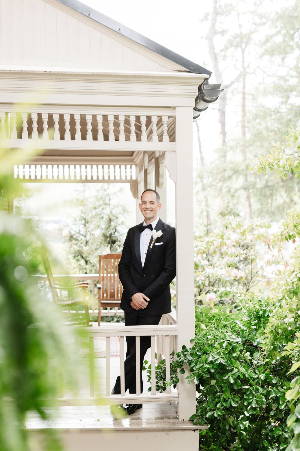 Smiling groom in black tuxedo with white boutonniere stands on white porch surrounded by lush green foliage.