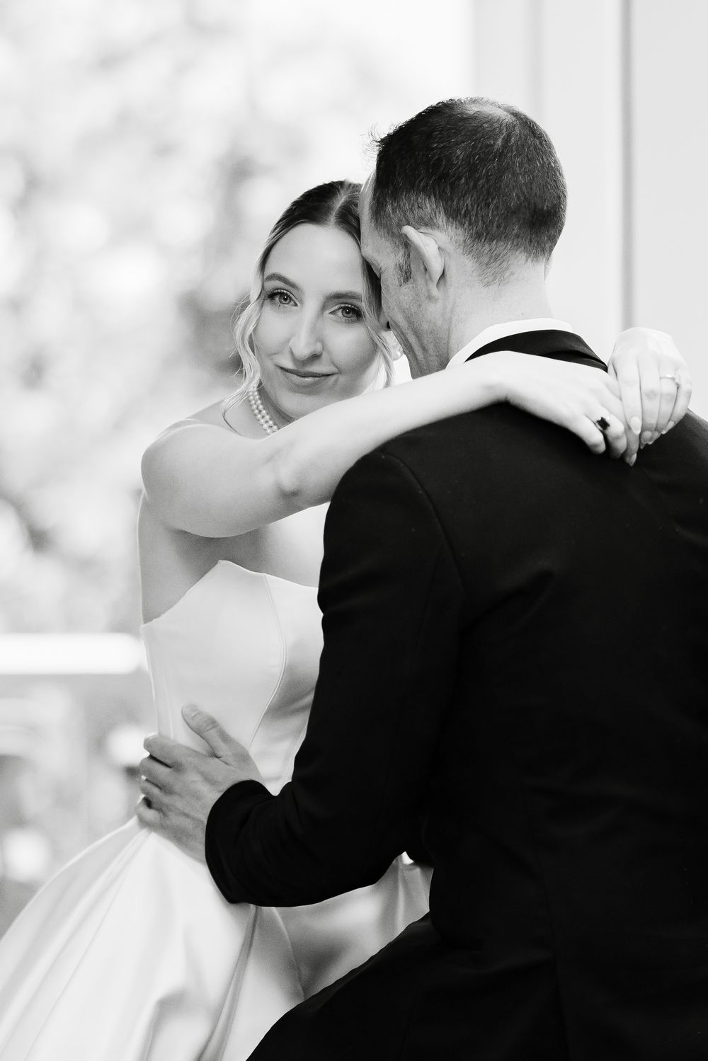 Bride in strapless white gown and pearl necklace embraces groom in black tuxedo in black and white photo.