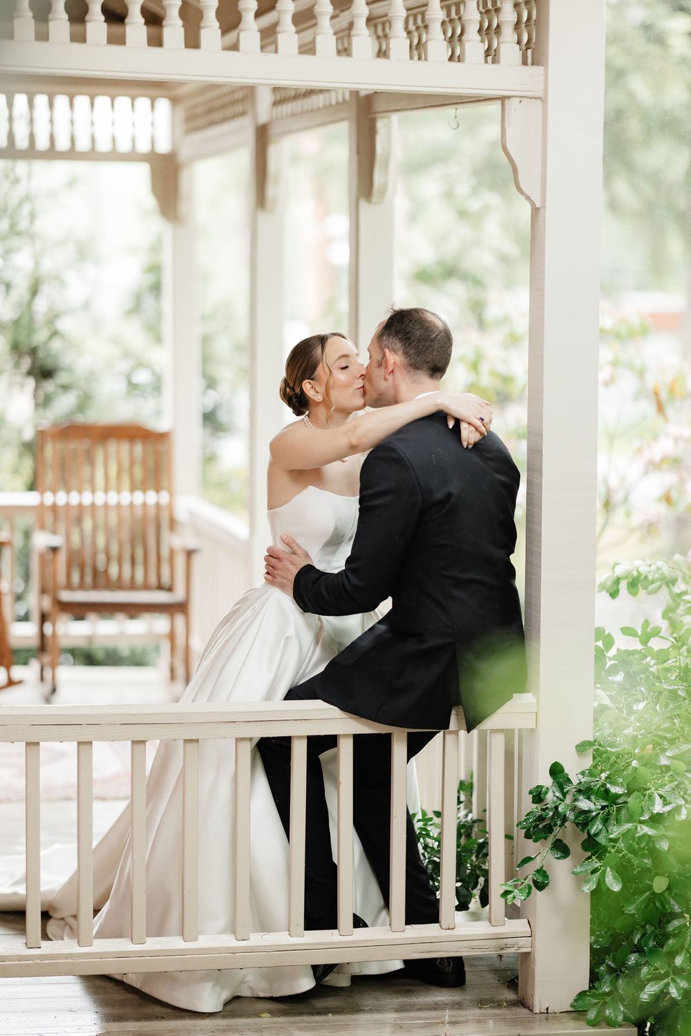 Bride and groom share a romantic kiss while embracing on a white decorative porch with garden backdrop.