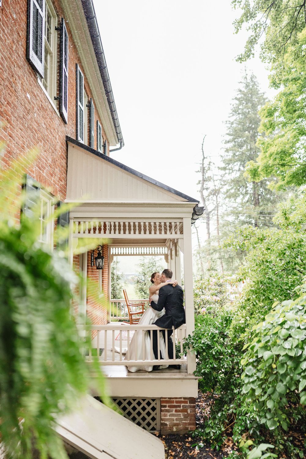 Bride and groom kiss on white Victorian porch beside brick building surrounded by lush green trees.