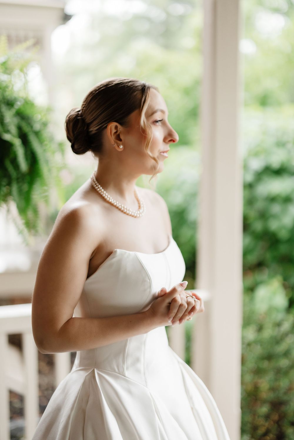 Elegant bride in strapless white gown and pearl necklace gazes serenely on a lush green porch.