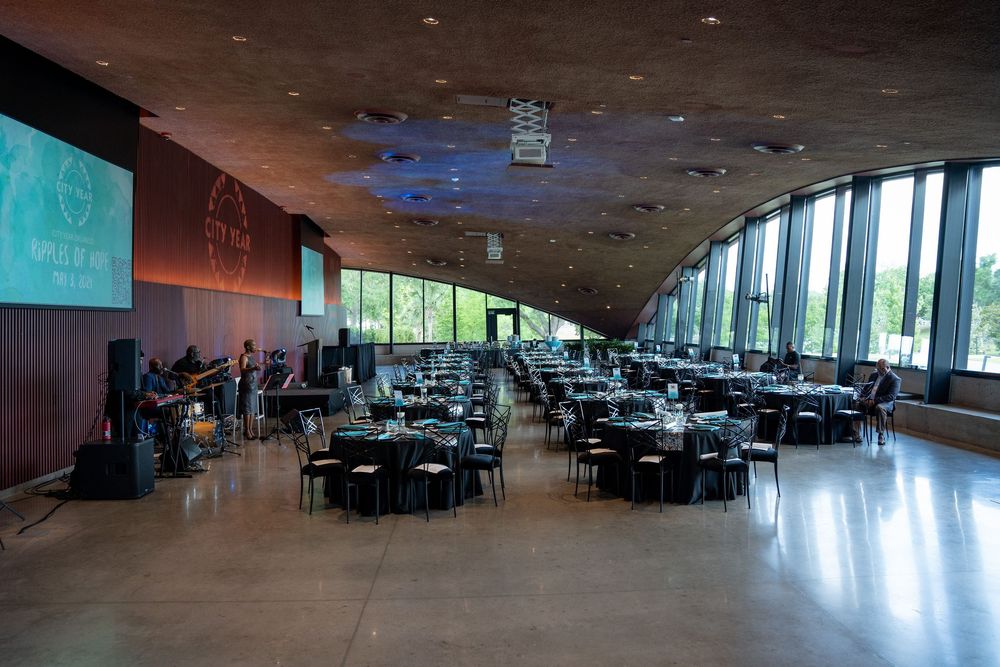 Elegant banquet hall with round tables, live band, large screen, and floor-to-ceiling windows overlooking greenery.