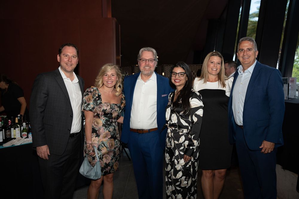 Six elegantly dressed guests smile together at an upscale indoor social event or gala dinner.