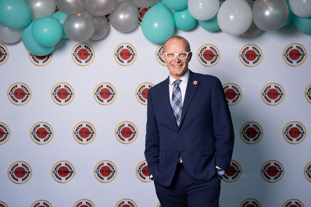 Man in navy suit smiling at event backdrop with balloons and branded logo wall.