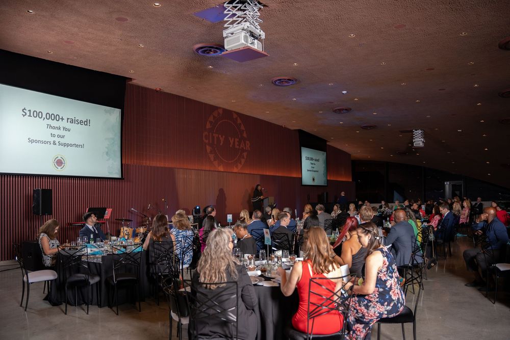 Elegant charity gala dinner event with guests seated at round tables beneath a large projection screen.