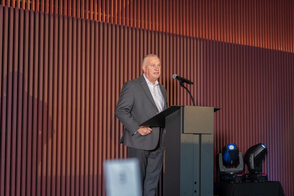 Man in gray suit speaking at podium during formal event with modern wood panel backdrop.