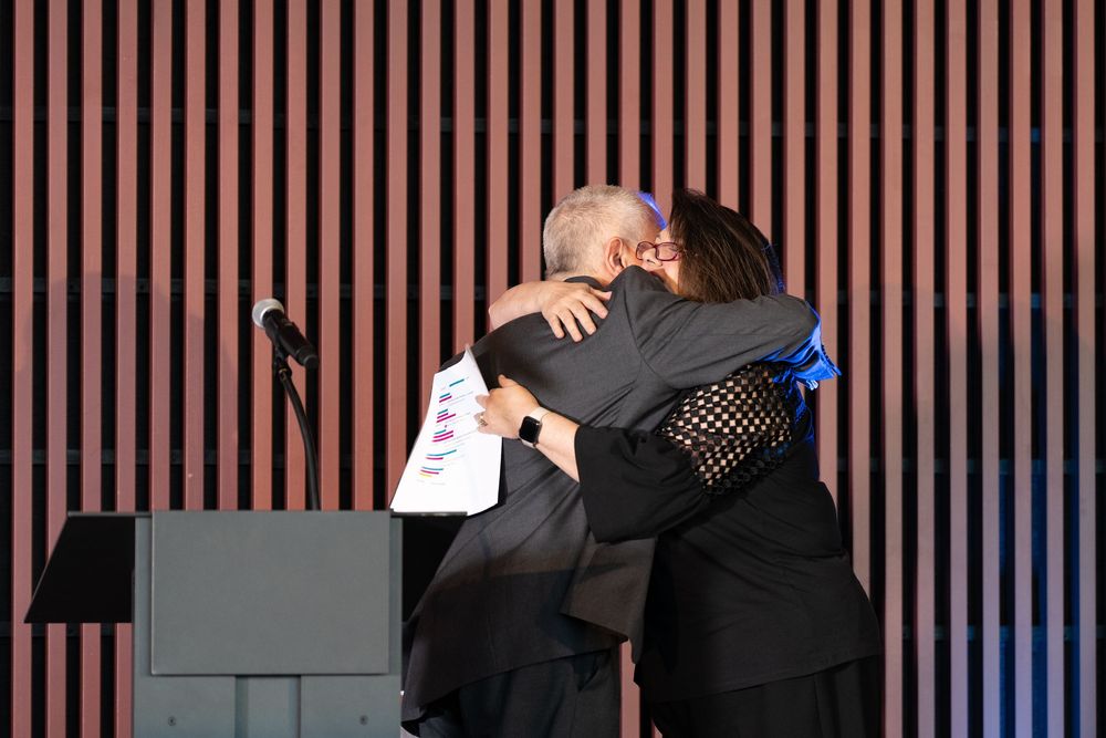 Two people share a warm embrace at a podium during a formal stage event.