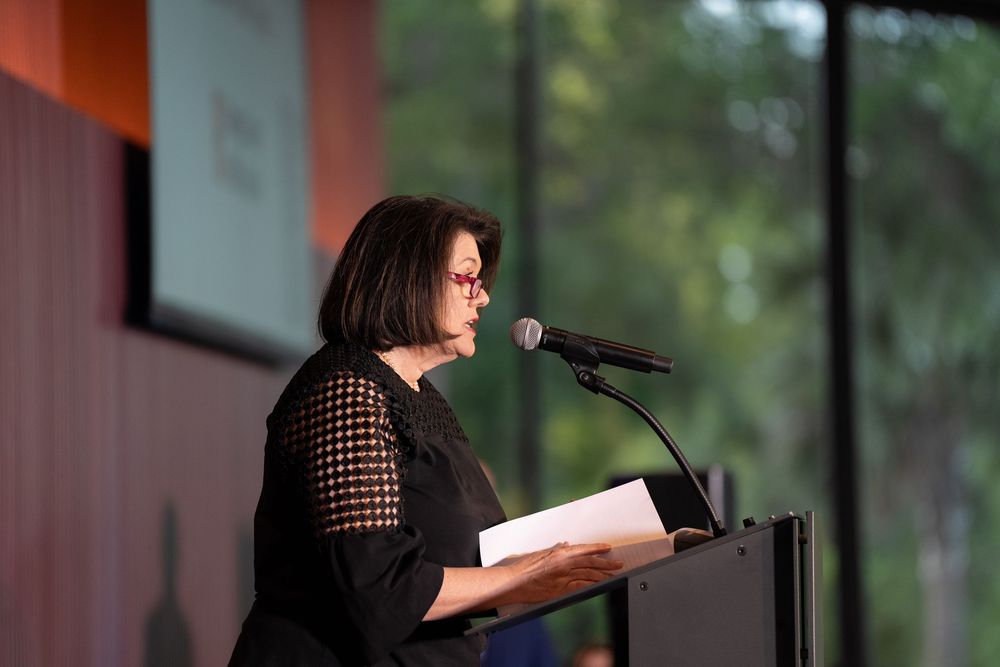 Woman in black outfit speaking at a podium with microphone during an indoor event or conference.