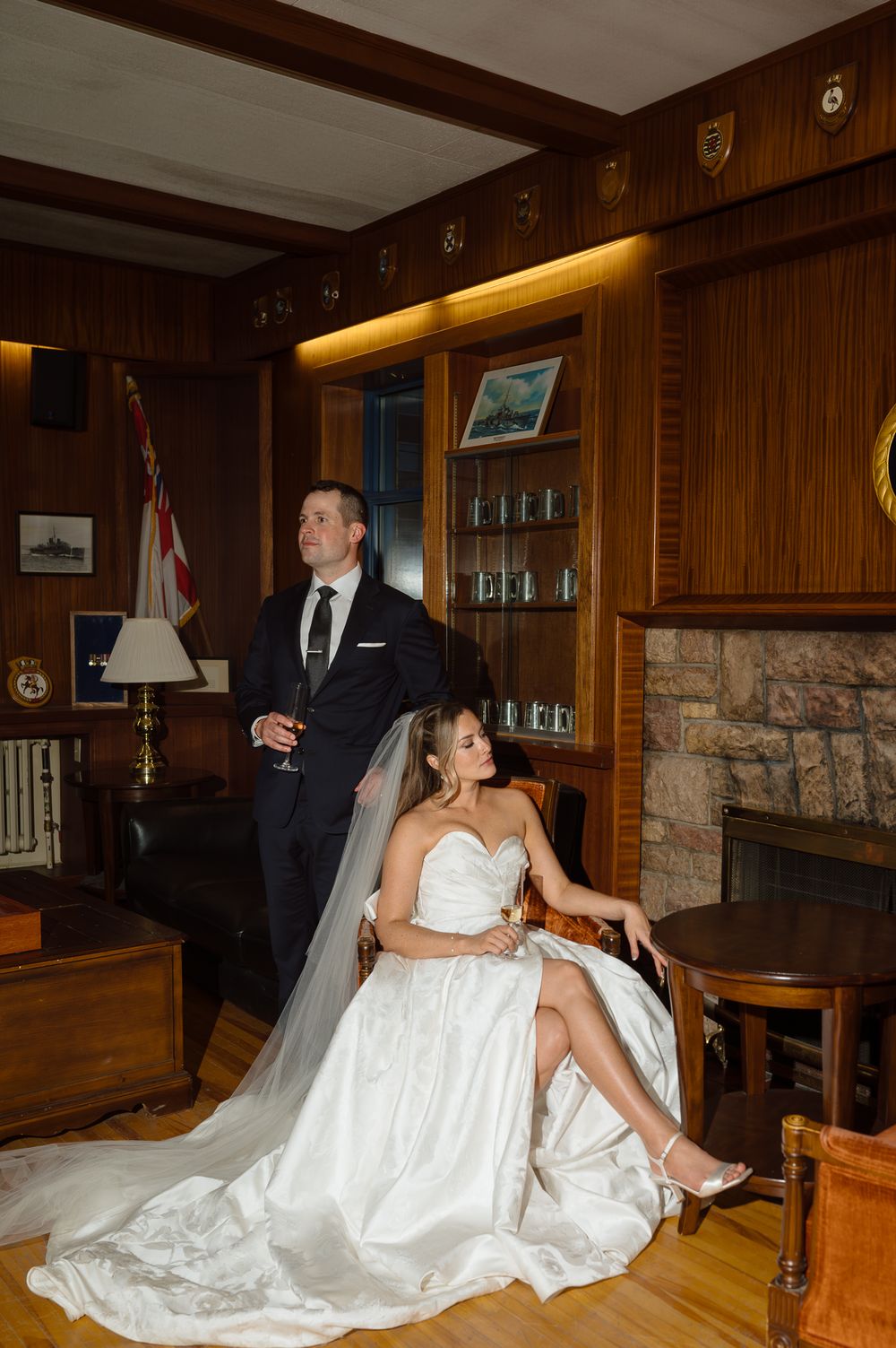 Bride in white gown seated beside groom in dark suit holding wine glass in elegant wood-paneled room with stone fireplace.