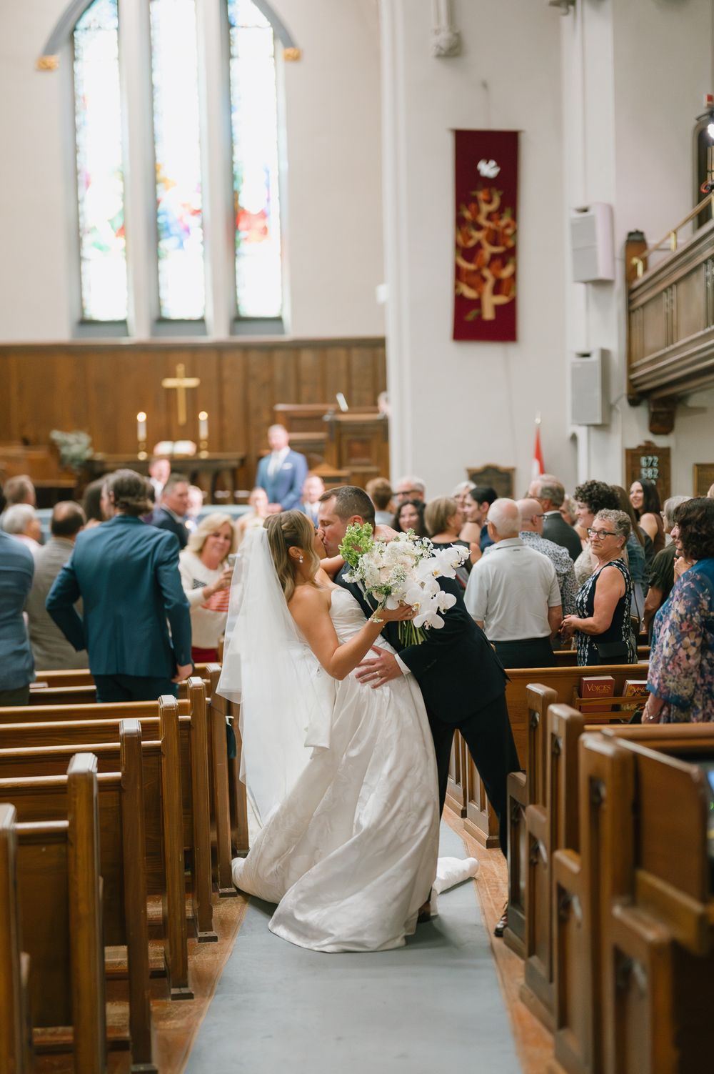 Bride walking down church aisle in white gown holding bouquet, guests standing in wooden pews during wedding ceremony.