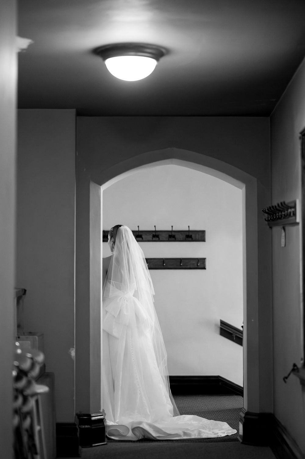Black and white photo of a bride in a flowing wedding gown and veil standing in an arched doorway.
