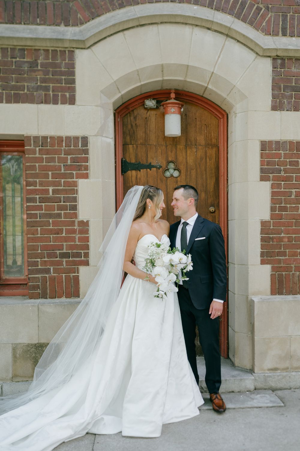 Bride in white gown with veil and groom in navy suit share a romantic moment outside a brick church.