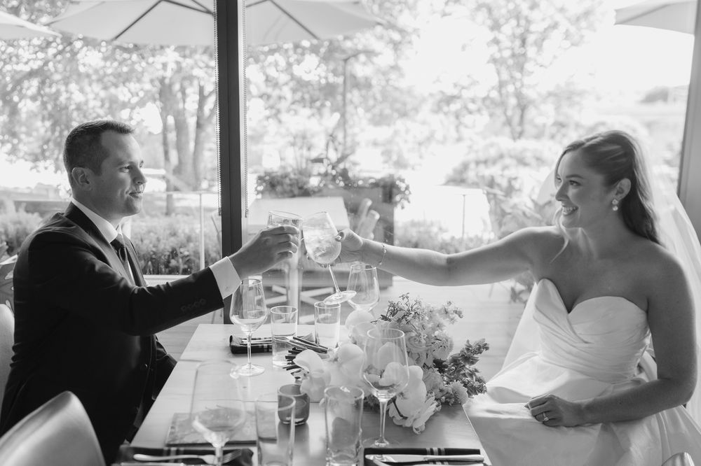 Bride and groom toasting with wine glasses at wedding reception table with floral centerpiece, black and white photo.