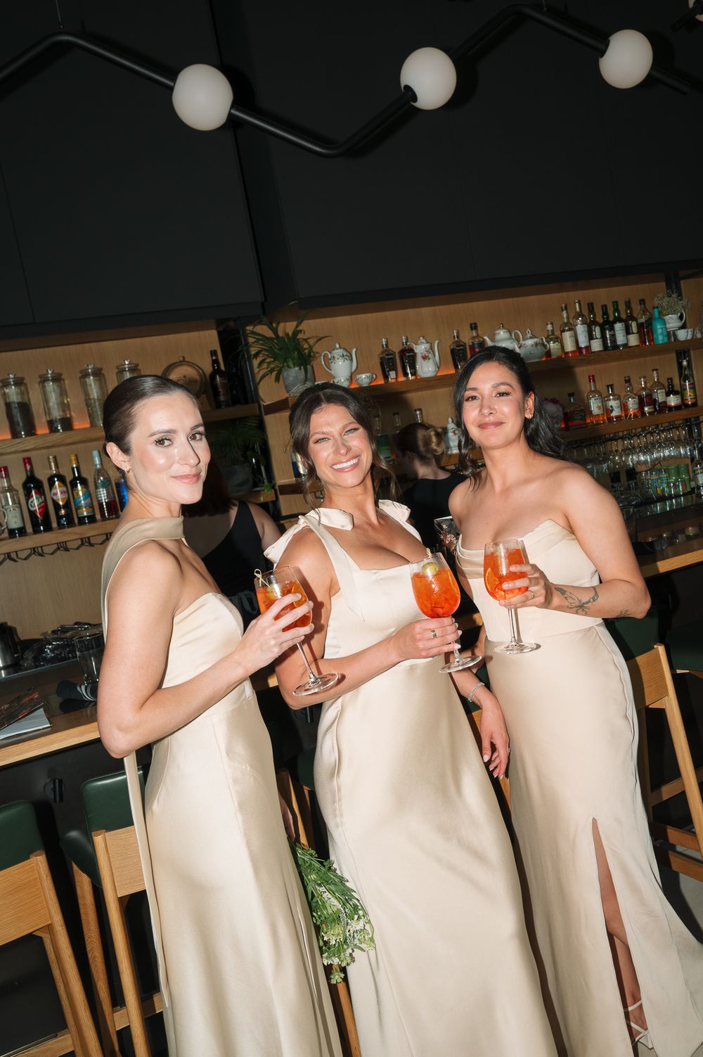 Three bridesmaids in champagne satin gowns holding Aperol Spritz cocktails at a bar venue.