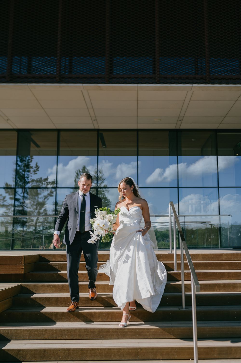 Bride and groom descend outdoor steps, bride holding white bouquet in elegant strapless gown on sunny wedding day.