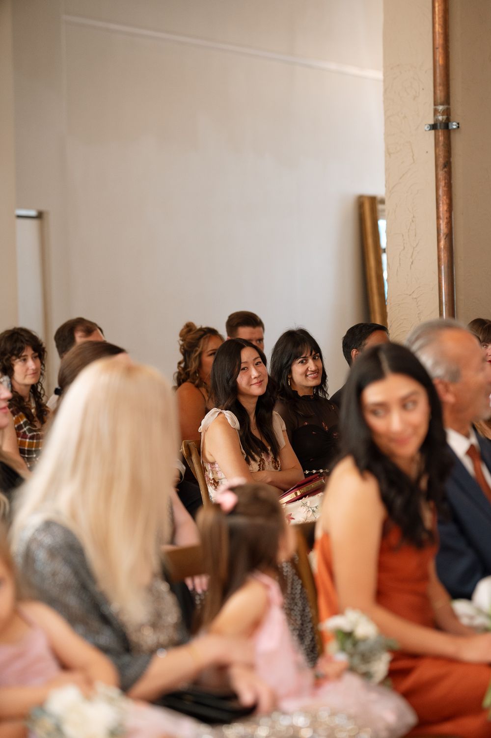 Smiling wedding guests sit in rows during a ceremony while a flower girl in a pink dress holds a bouquet in the foreground.