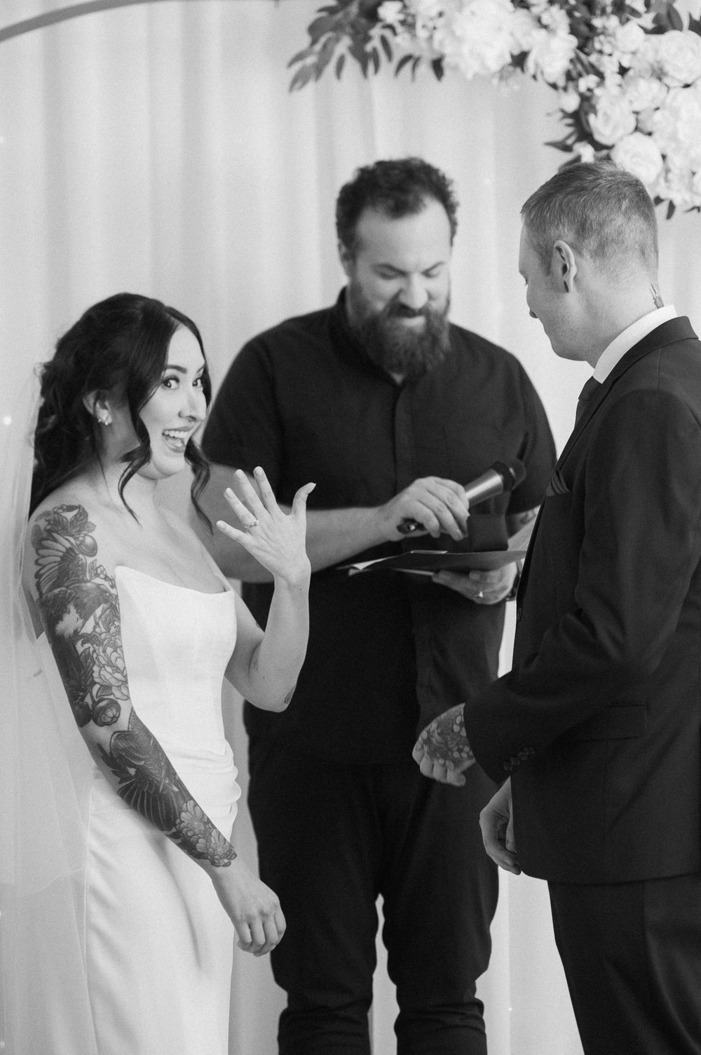 Smiling tattooed bride in a strapless gown holds up her hand as an officiant reads vows during a black and white wedding ceremony.