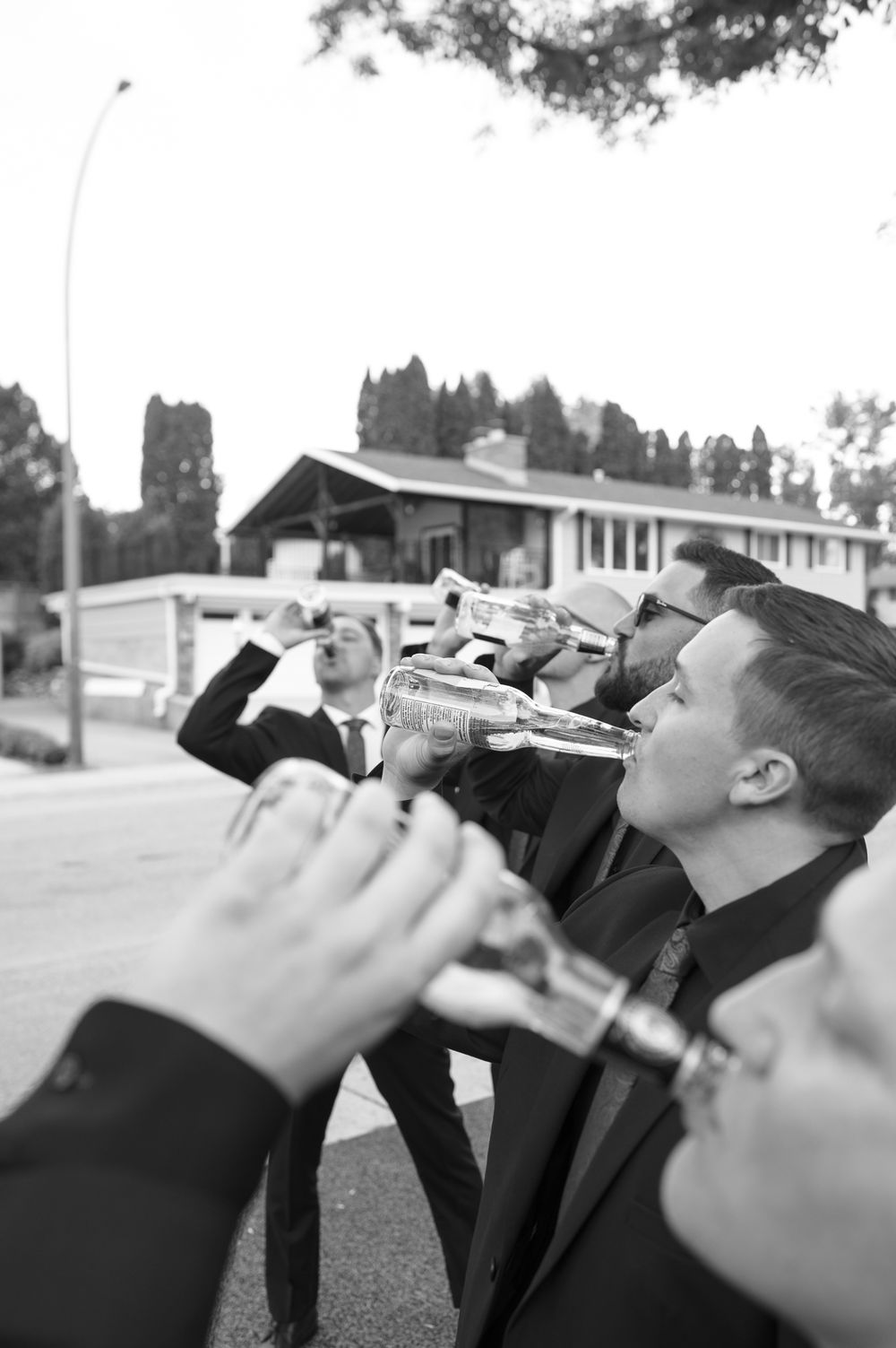 Black and white photo of groomsmen in suits drinking from bottles outdoors near a building.
