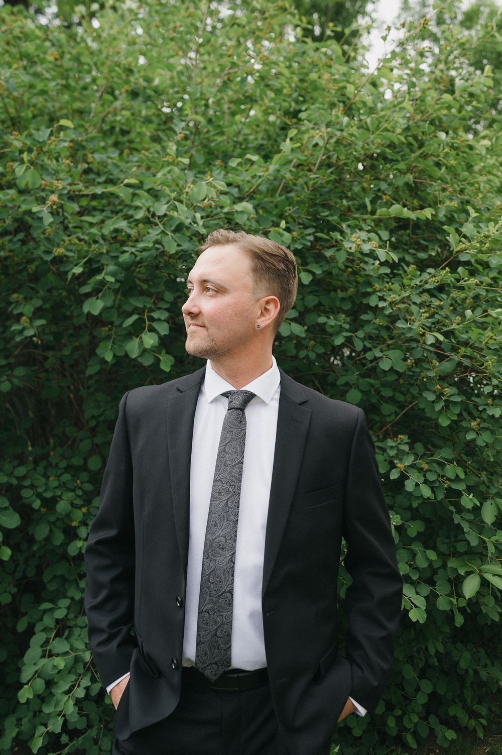 A man in a black suit and gray tie stands confidently with hands in pockets before lush green bushes.