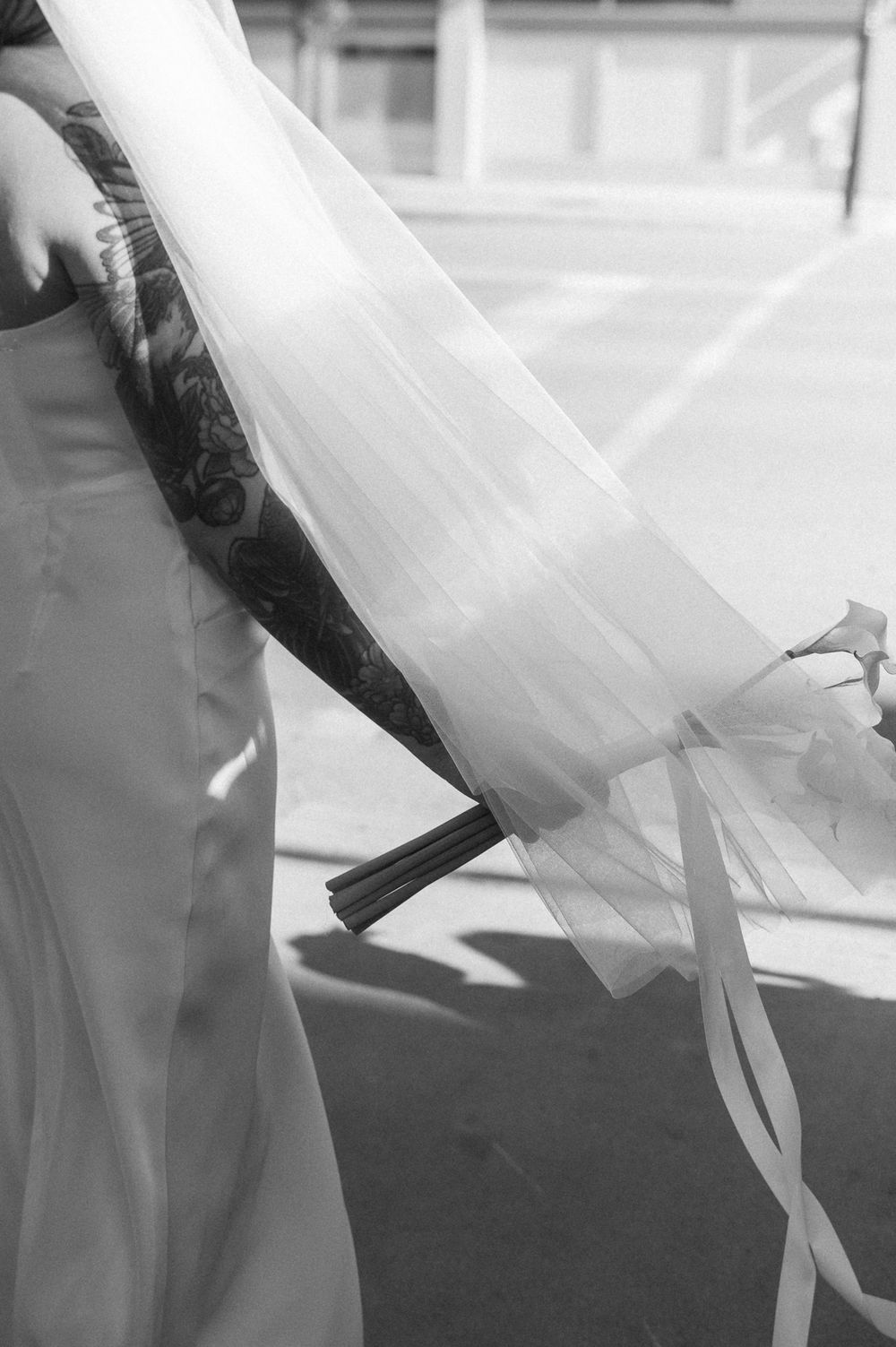 Black and white photo of a bride in a strapless gown with a sheer veil and tattooed arm holding a bouquet with trailing ribbons.