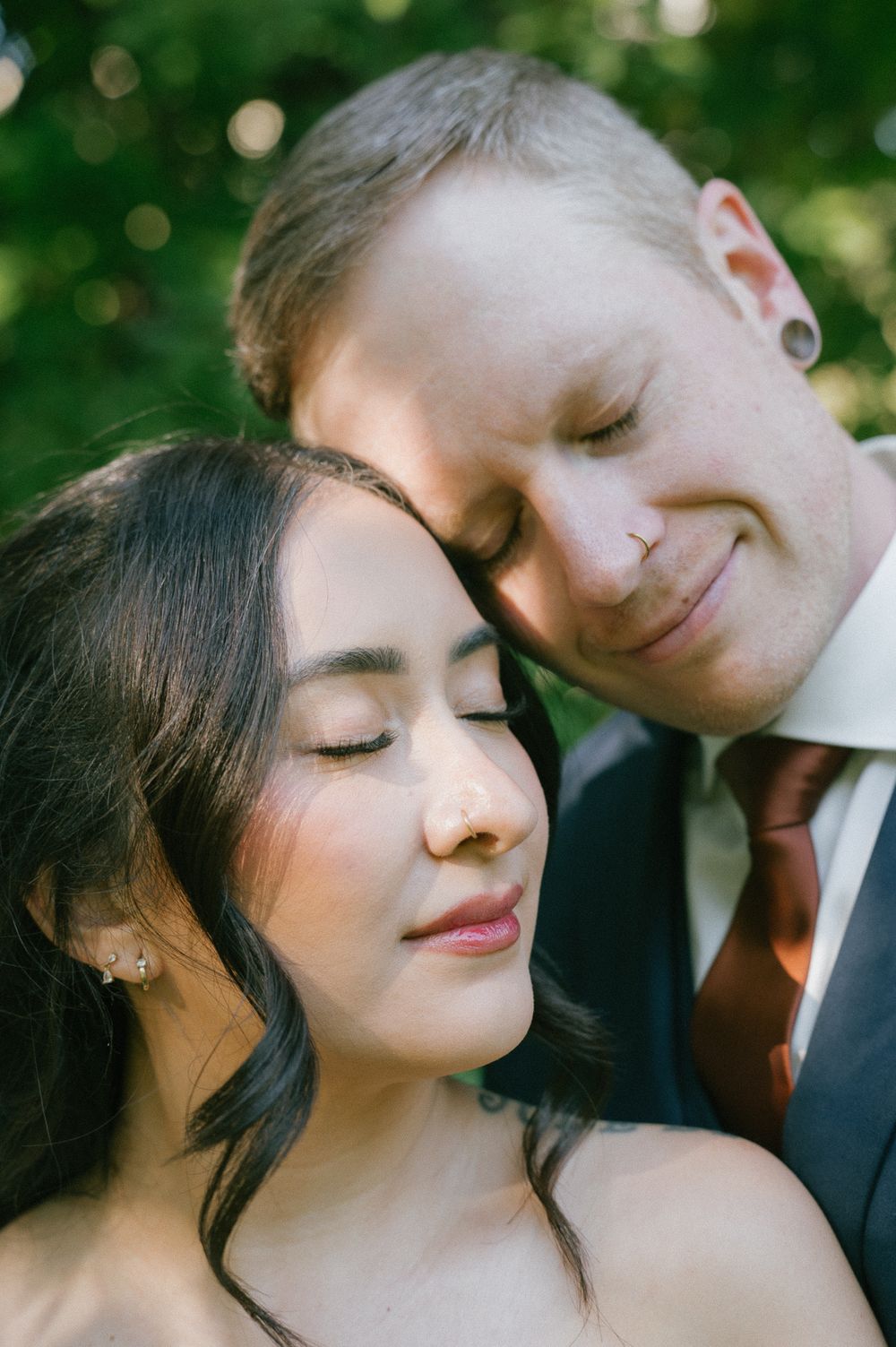 Smiling man in a suit leans his head against a woman with closed eyes and dark hair in a tender embrace outdoors among green foliage.