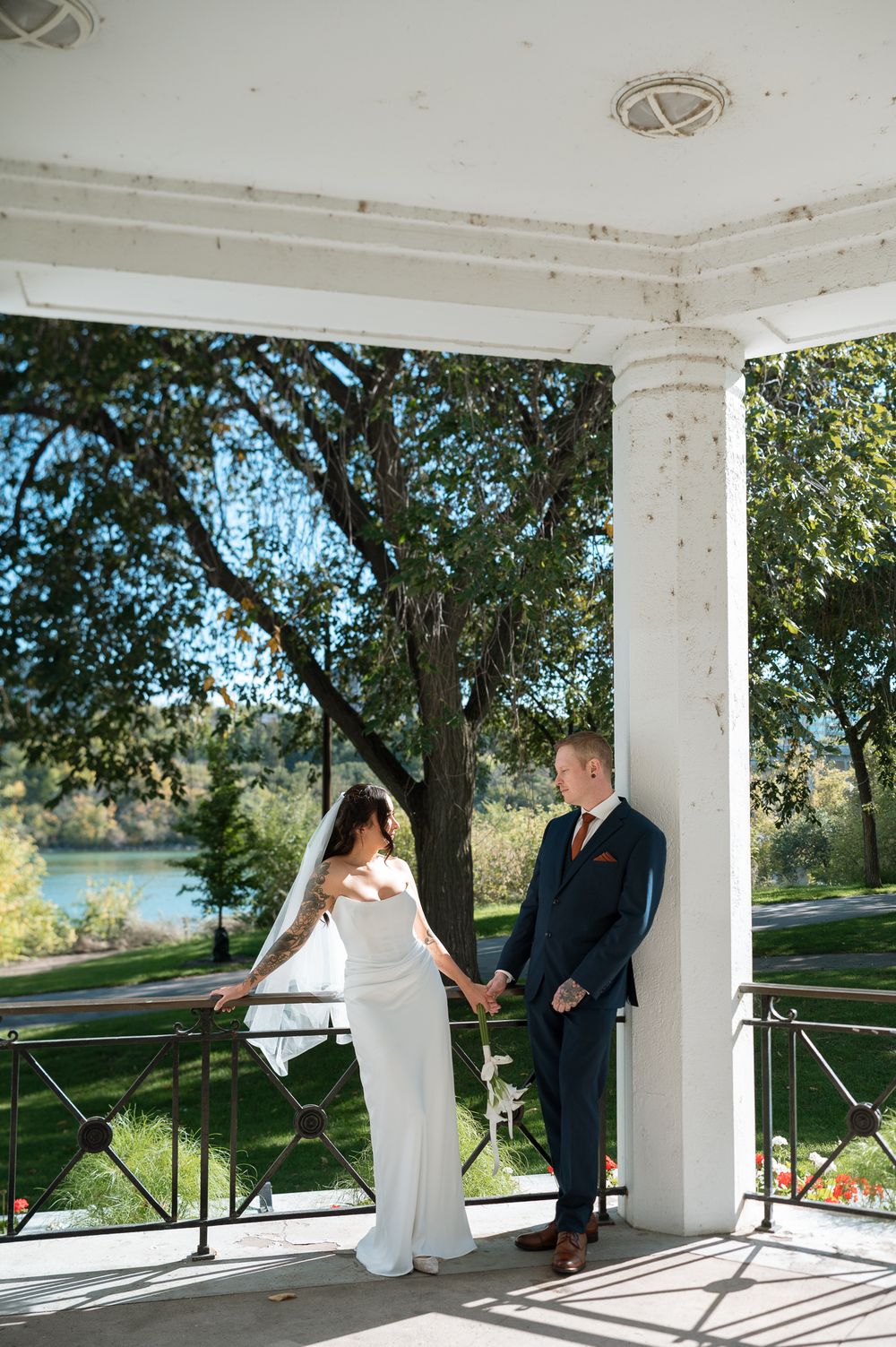 Bride in a white gown and veil holds hands with a groom in a navy suit under a white pavilion overlooking a park and river.