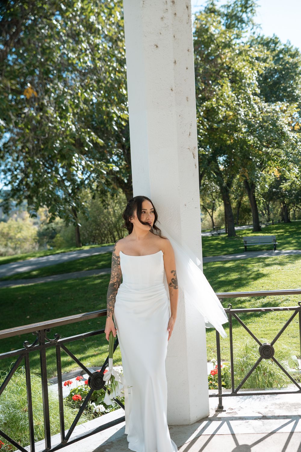 Bride with arm tattoos in a strapless white wedding gown and veil poses beside a white column on a sunny balcony overlooking green trees.