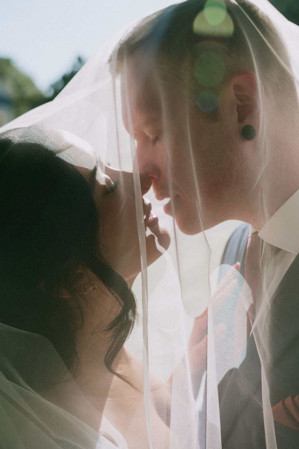 A bride and groom share a tender kiss beneath a sheer wedding veil, with warm sunlight and soft bokeh glowing around them.