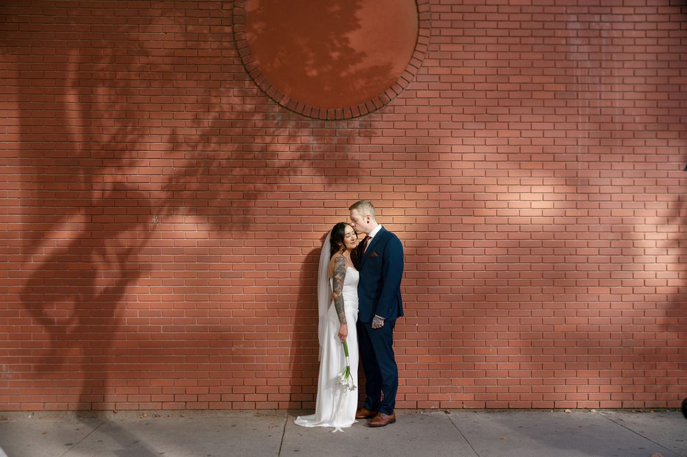 Bride in a white gown and groom in a navy suit embrace in front of a red brick wall with soft tree shadows and a round medallion.