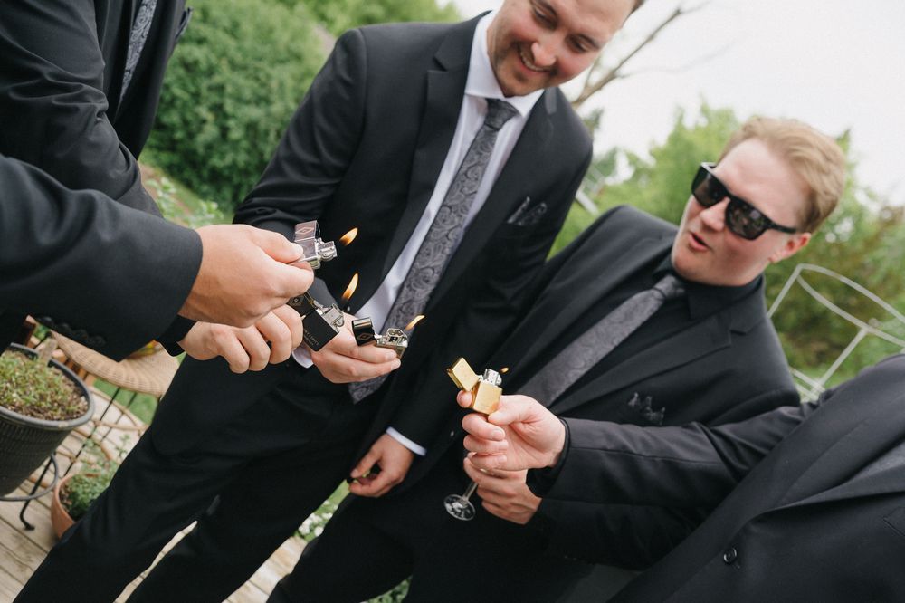 Three men in black suits lighting cigars outdoors, one wearing sunglasses, at a formal celebration.