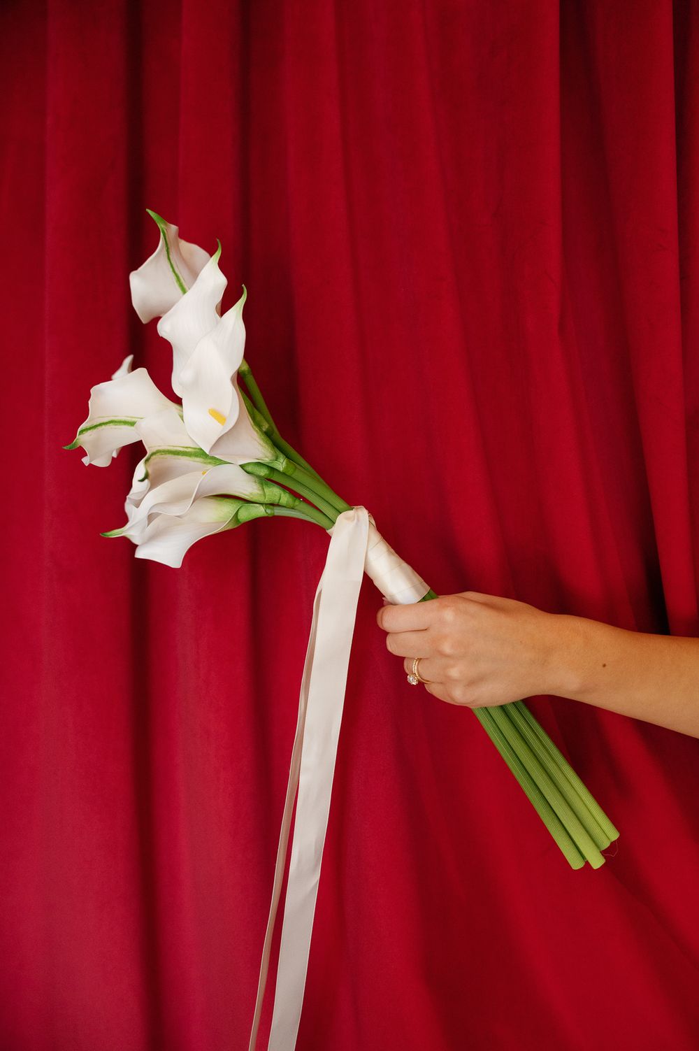 Hand holding a bridal bouquet of white calla lilies with long green stems and a cream ribbon against a red velvet curtain.
