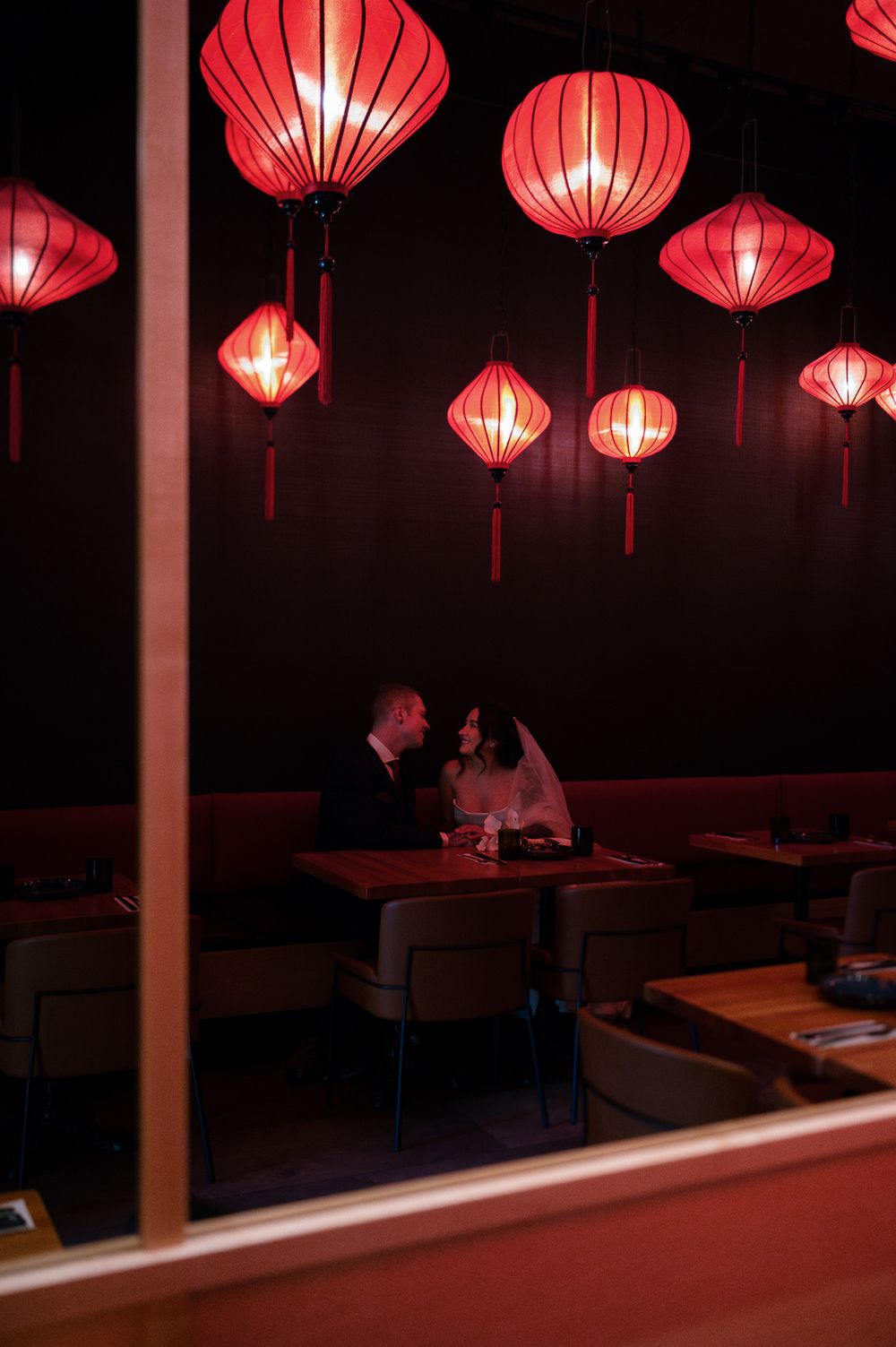 Bride and groom share an intimate moment in a dim restaurant booth beneath glowing red silk lanterns, seen through a window.