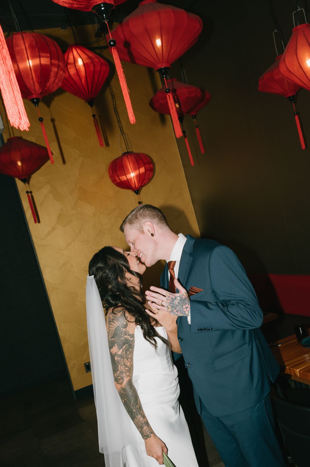 Bride in a white gown and veil kisses her groom in a blue suit beneath glowing red paper lanterns at a moody wedding reception.