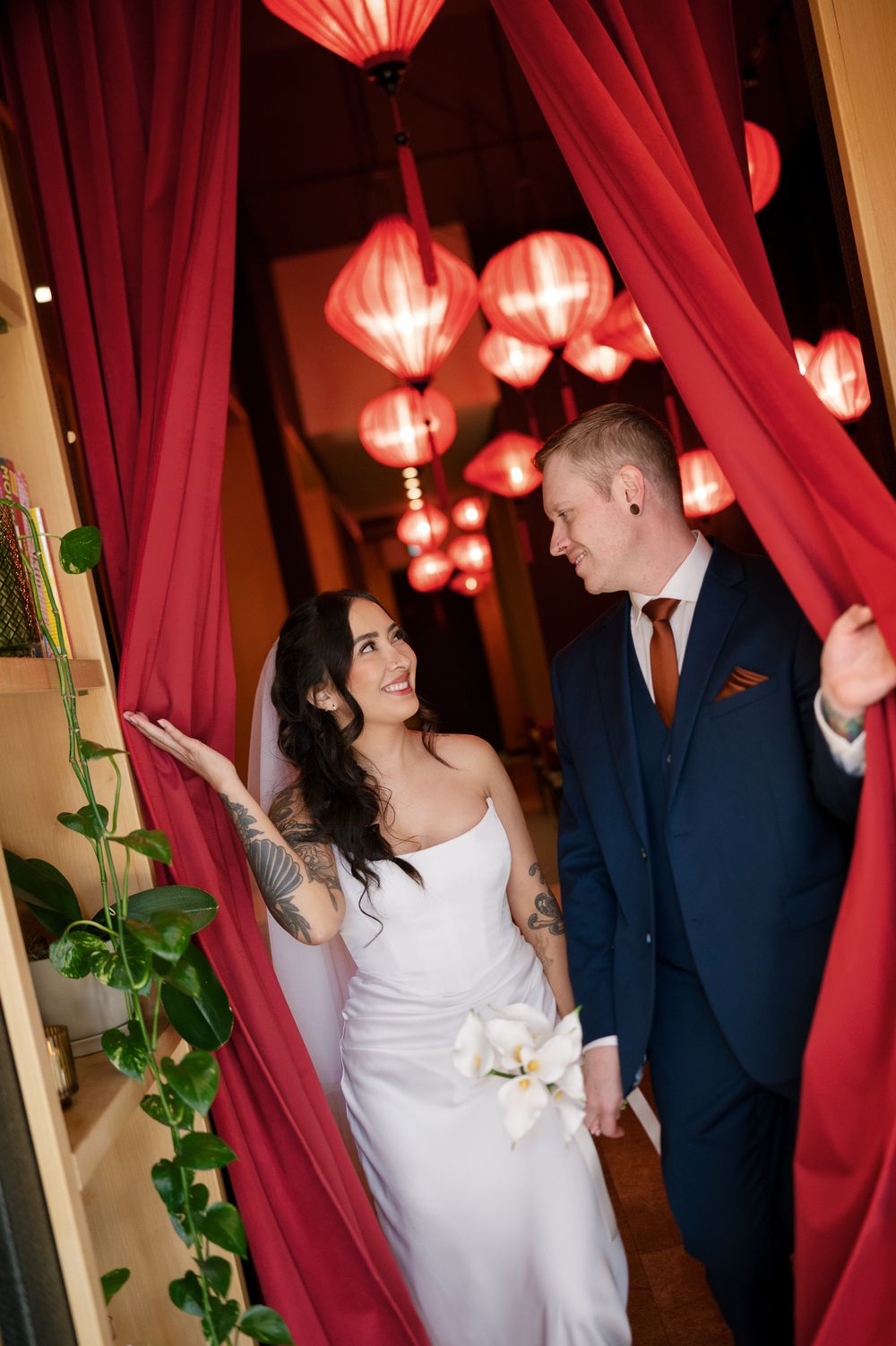 Bride in a white strapless gown and groom in a navy suit smile at each other between red curtains beneath glowing red paper lanterns.