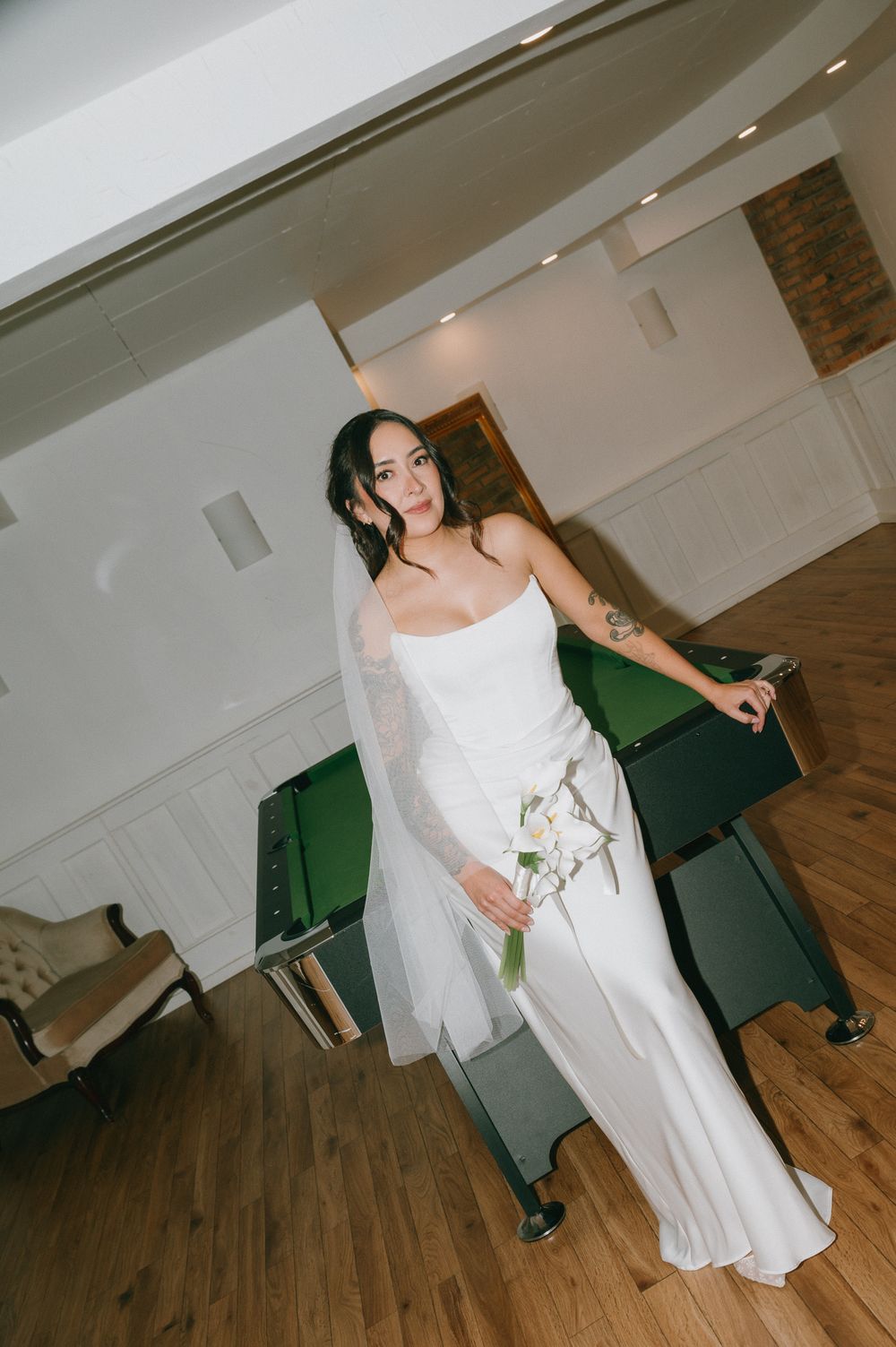 Bride in a white strapless satin gown and sheer veil holding a white floral bouquet beside a green pool table in a bright room.