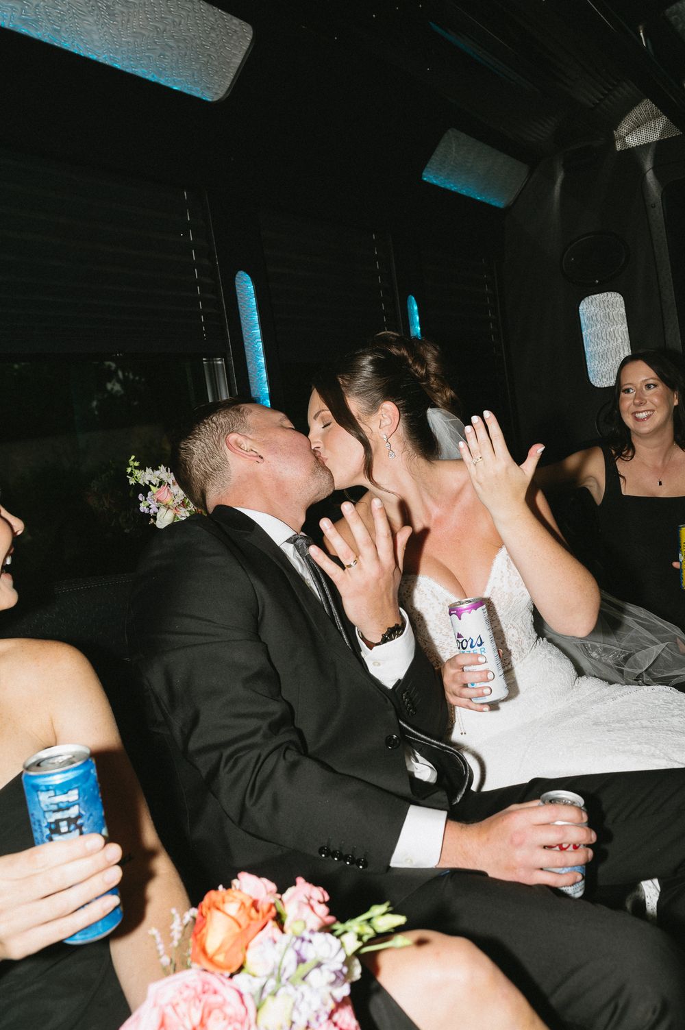 Bride and groom share a kiss in a party bus while holding canned drinks, with bridesmaids smiling nearby.
