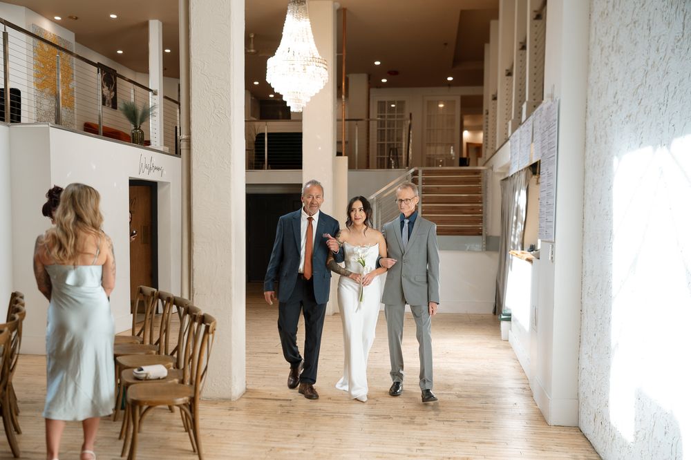 Bride in a white gown walks arm in arm with two men in suits through a bright, elegant wedding venue with a chandelier and wooden chairs.