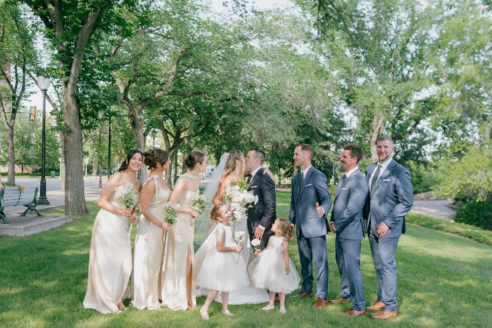 Wedding party poses outdoors on green lawn, bridesmaids in champagne gowns, groomsmen in gray suits, with flower girls in white dresses.