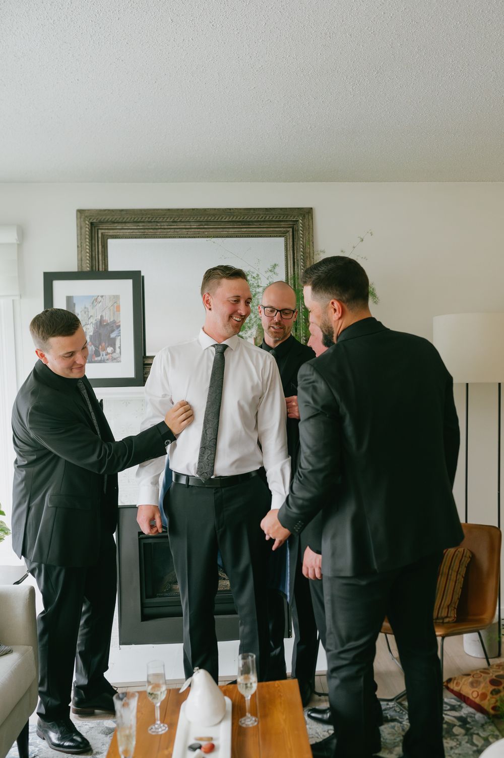 Groomsmen helping groom get dressed in white shirt and grey tie before wedding ceremony indoors.