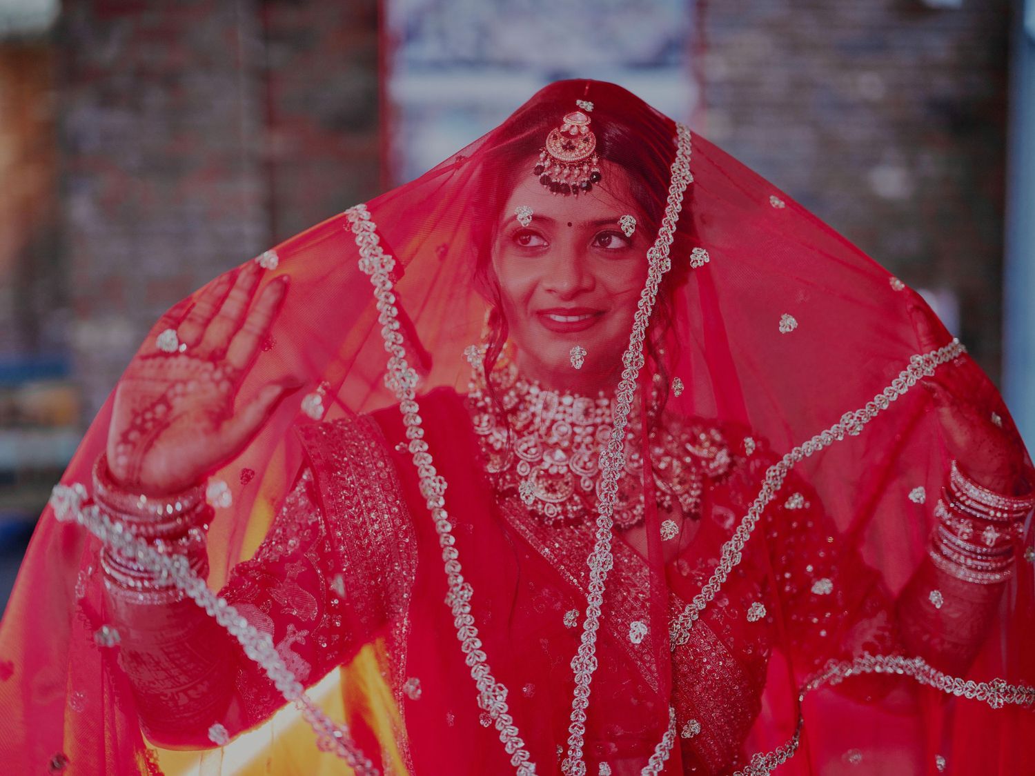 A bride in a red bridal lehenga and dupatta with intricate silver embroidery, wearing traditional jewelry, waving her mehndi-adorned hand.