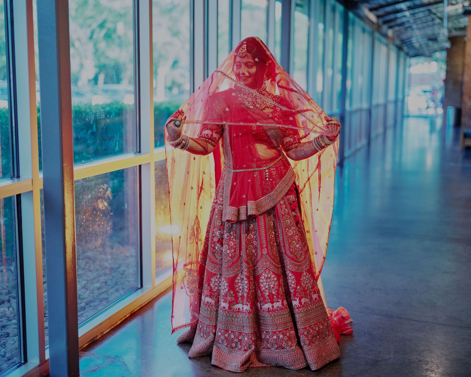 Indian bride in red lehenga choli with dupatta posing elegantly in a sunlit corridor with large windows.