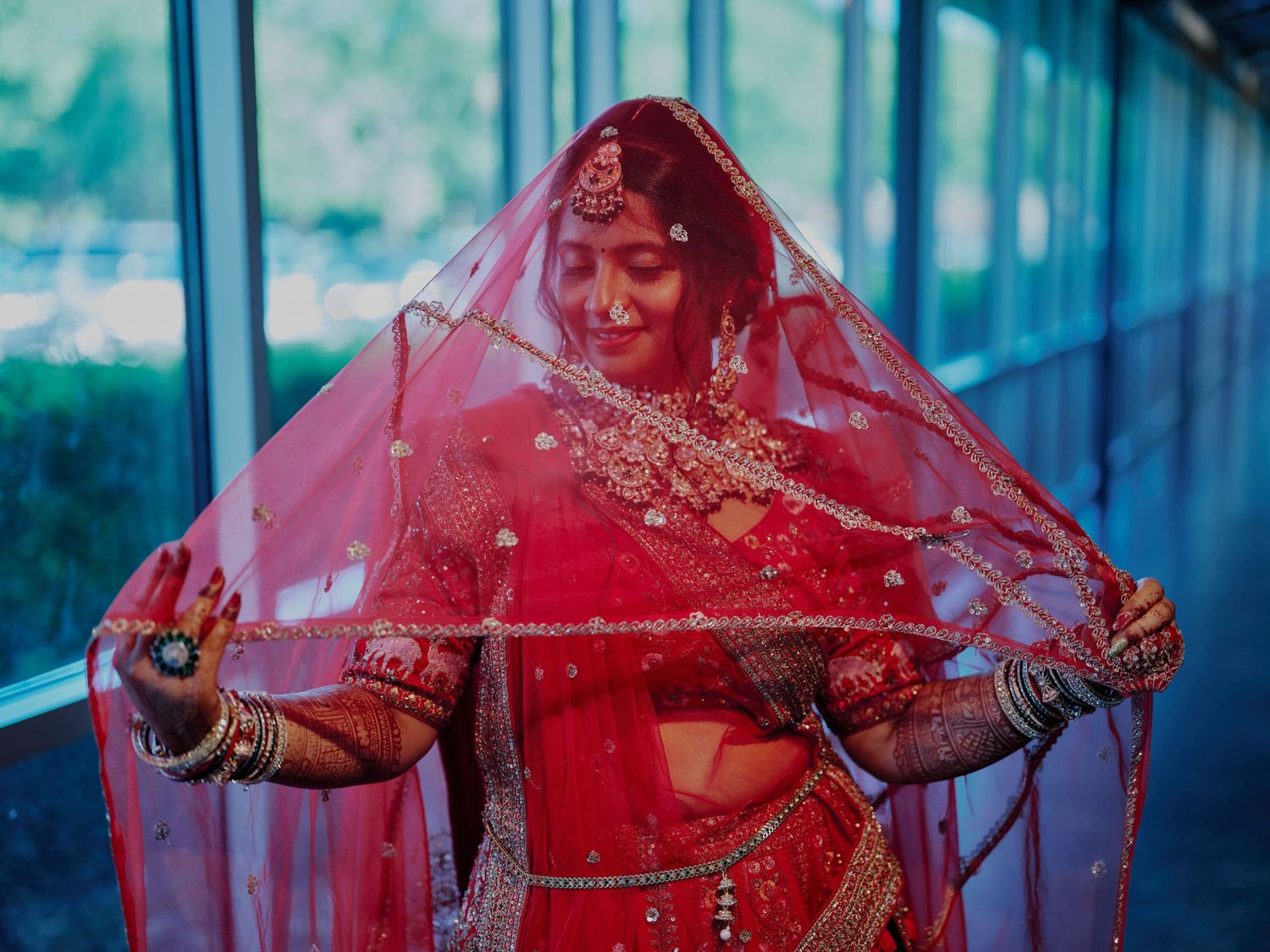 Indian bride in red lehenga and dupatta showcasing bridal jewelry and mehndi at a wedding ceremony.