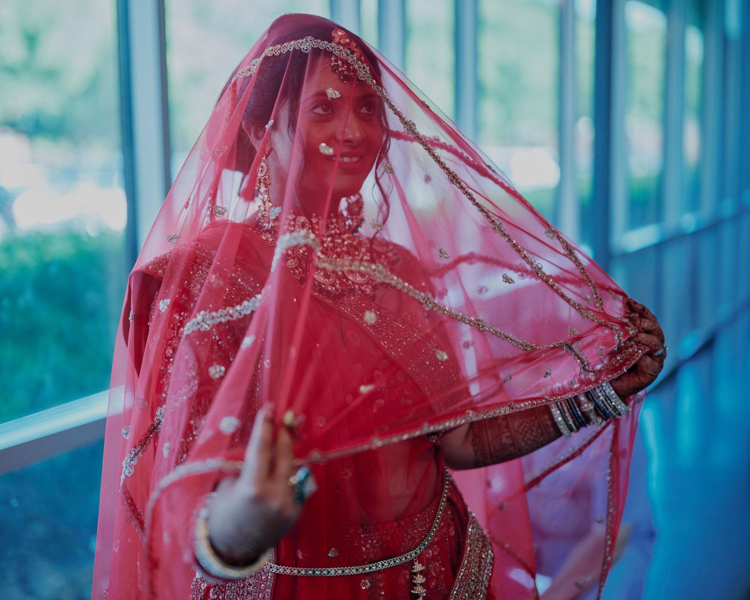 Indian bride in red bridal lehenga and dupatta with gold jewelry, posing gracefully at her wedding ceremony.